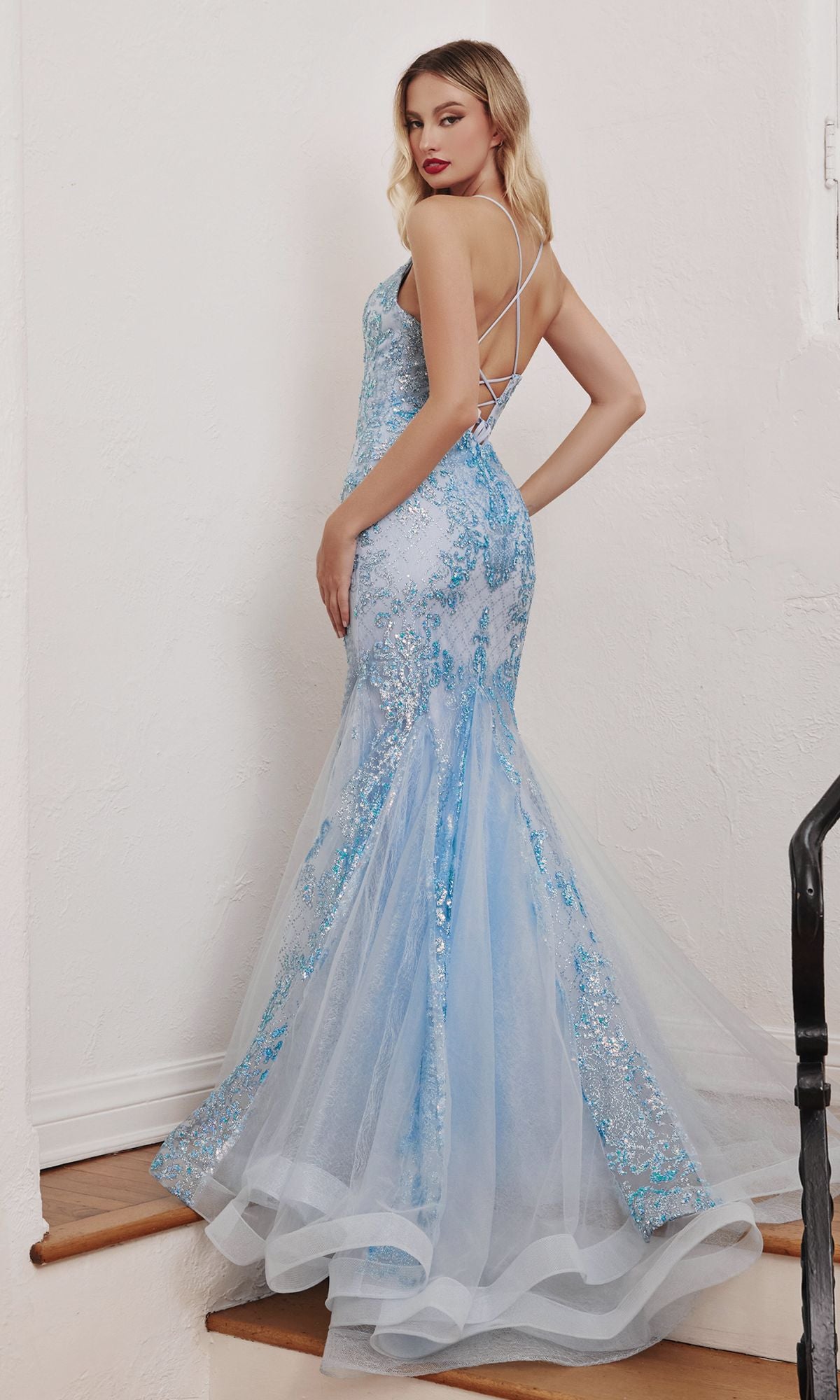 A woman faces away on stairs, wearing the Strappy-Back Long Beaded Mermaid Prom Dress CC2279, a light blue floor-length gown with sequins and thin straps.
